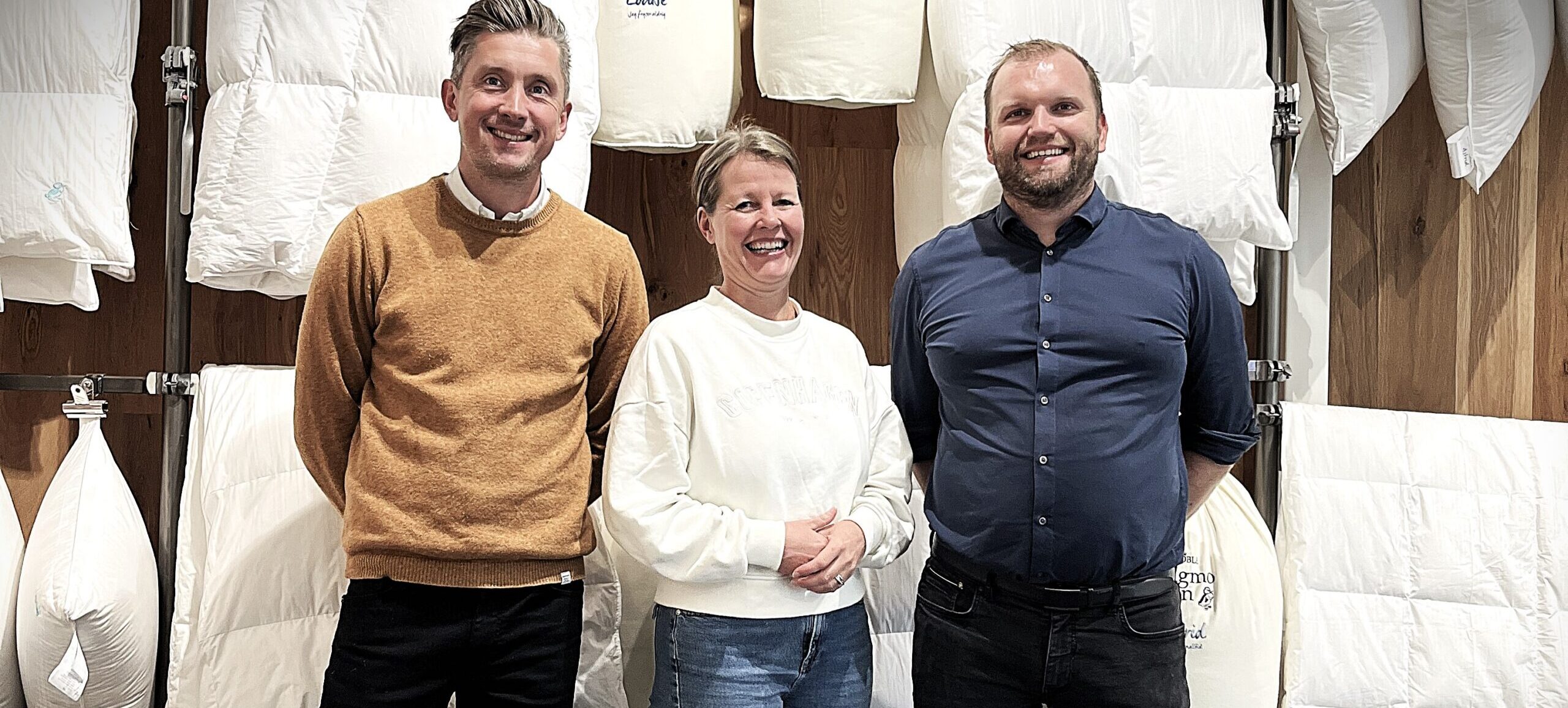 Tre personer står foran en væg fyldt med hvide dyner og puder, der er udstillet i et showroom.