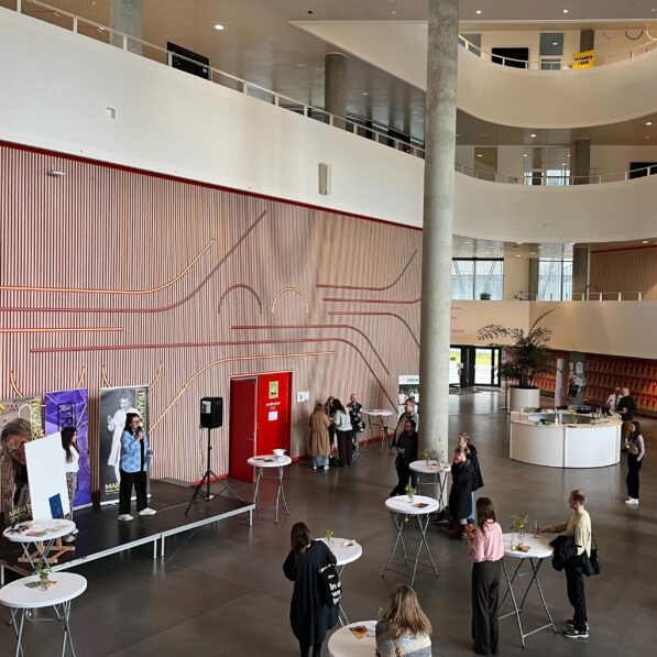 Atriet er et stort åbent rum ved SDU Koldings hovedindgang. Det er fyldt med mennesker, der står ved spredte runde borde og kigger op mod en scene, hvor to kvinder fra Trapholt præsenterer en ny kunstudstilling.