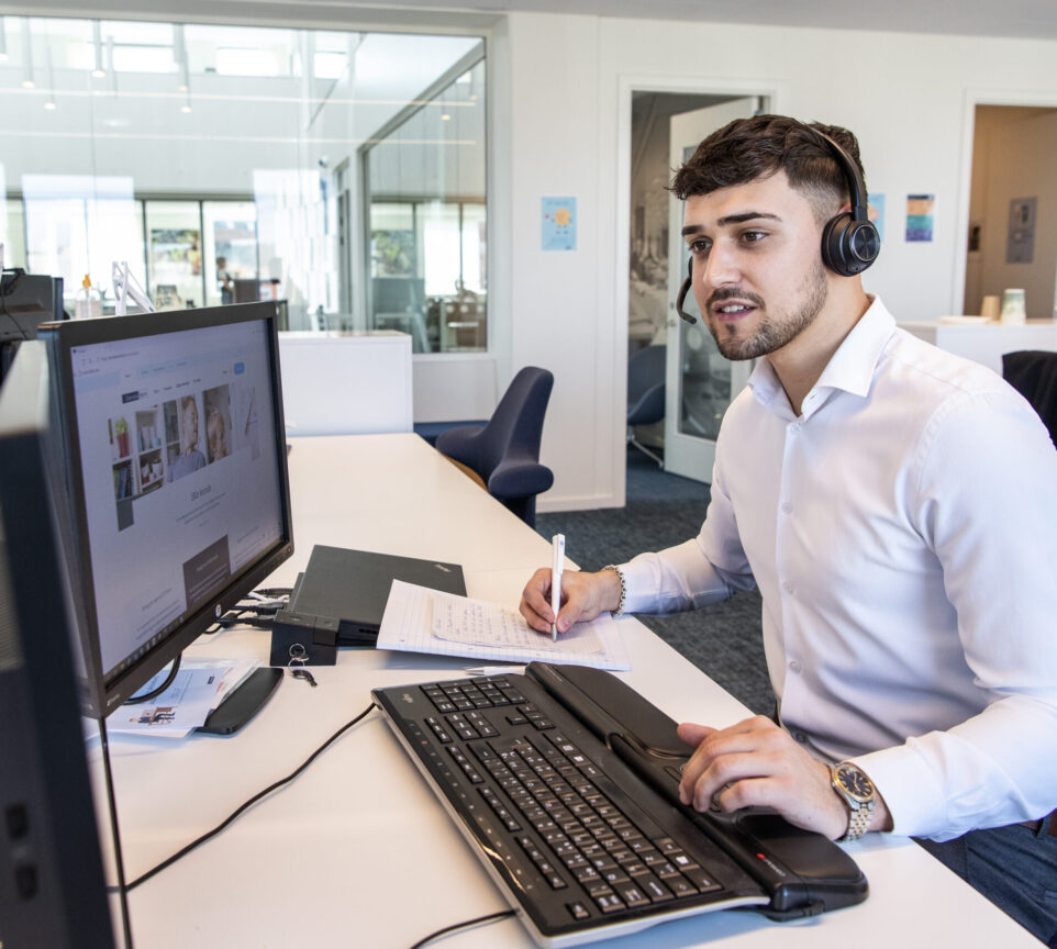Mergim sidder ved et skrivebord. Han er iført headset og kigger på en computerskærm, imens han skriver noget på en blok.