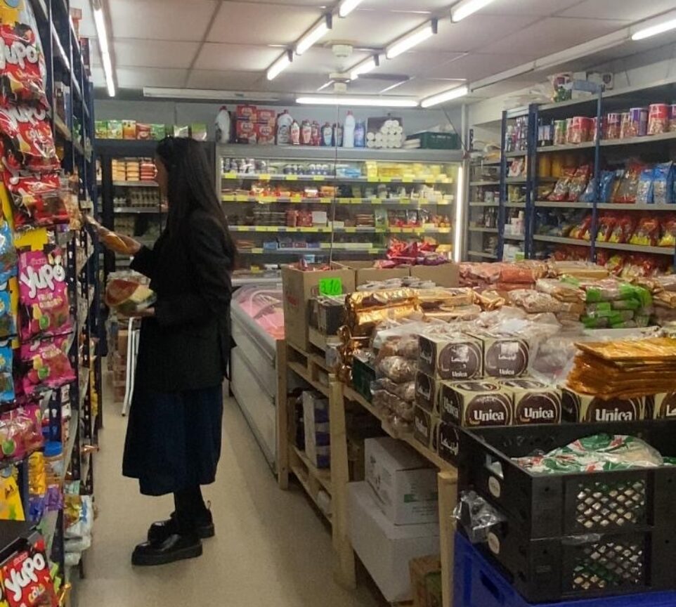 Sara Bilić står i en dagligdagsbutik, hvor hun er omgivet af hylder fyldt med madvarer.