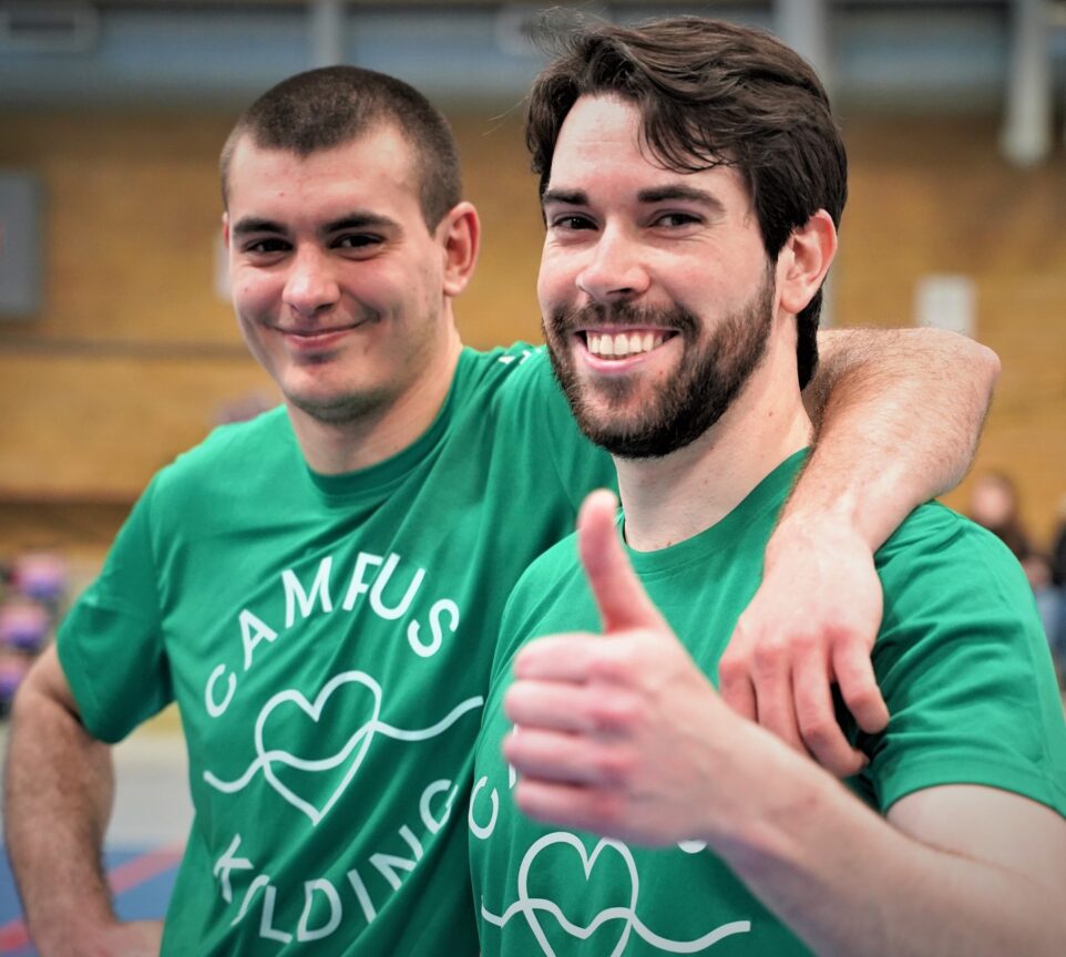 To unge mænd smiler mod kameraet, iført grønne Campus Kolding T-shirts.