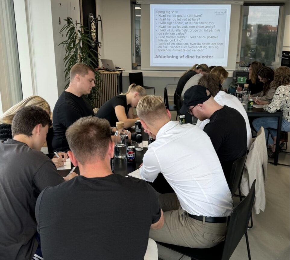 En gruppe unge mennesker sidder omkring 2 bord og skriver notater. Der er en skærm med en PowerPoint præsentation i baggrunden.