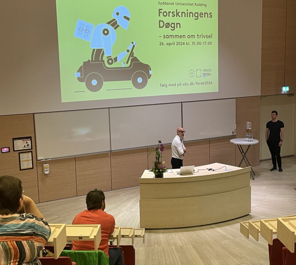 Mikael Sorknæs er iført jakkesæt og præsenterer temaet for Forskningens Døgn foran et publikum og et lærred med teksten "Forskningens Døgn - sammen om trivsel".