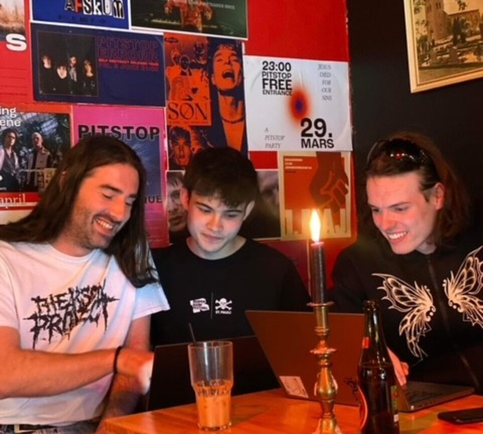 3 unge mænd sidder i hjørnet af en cafe og kigger smilende på en PC.